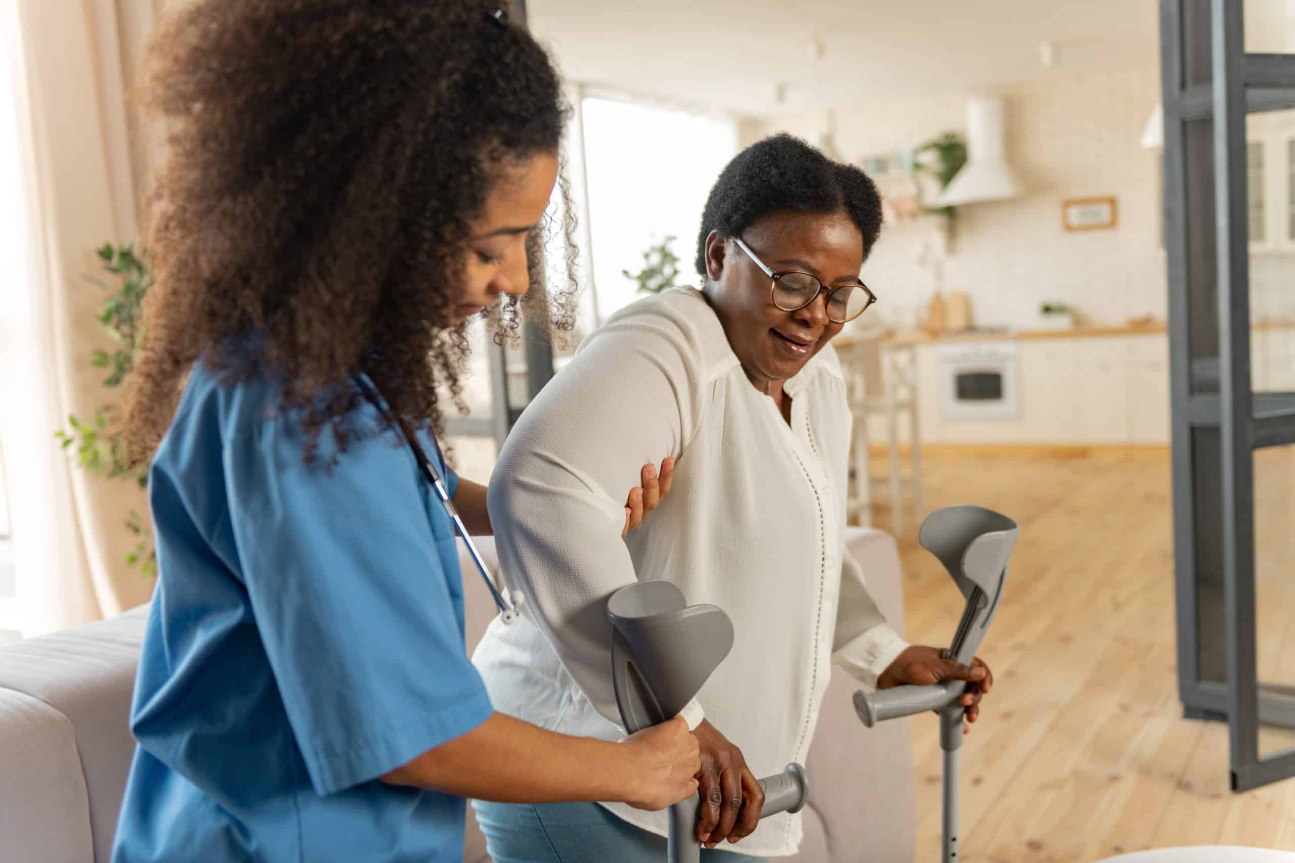 Giving crutches. Curly young dark-haired nurse giving crutches to aged woman wearing glasses after leg surgery